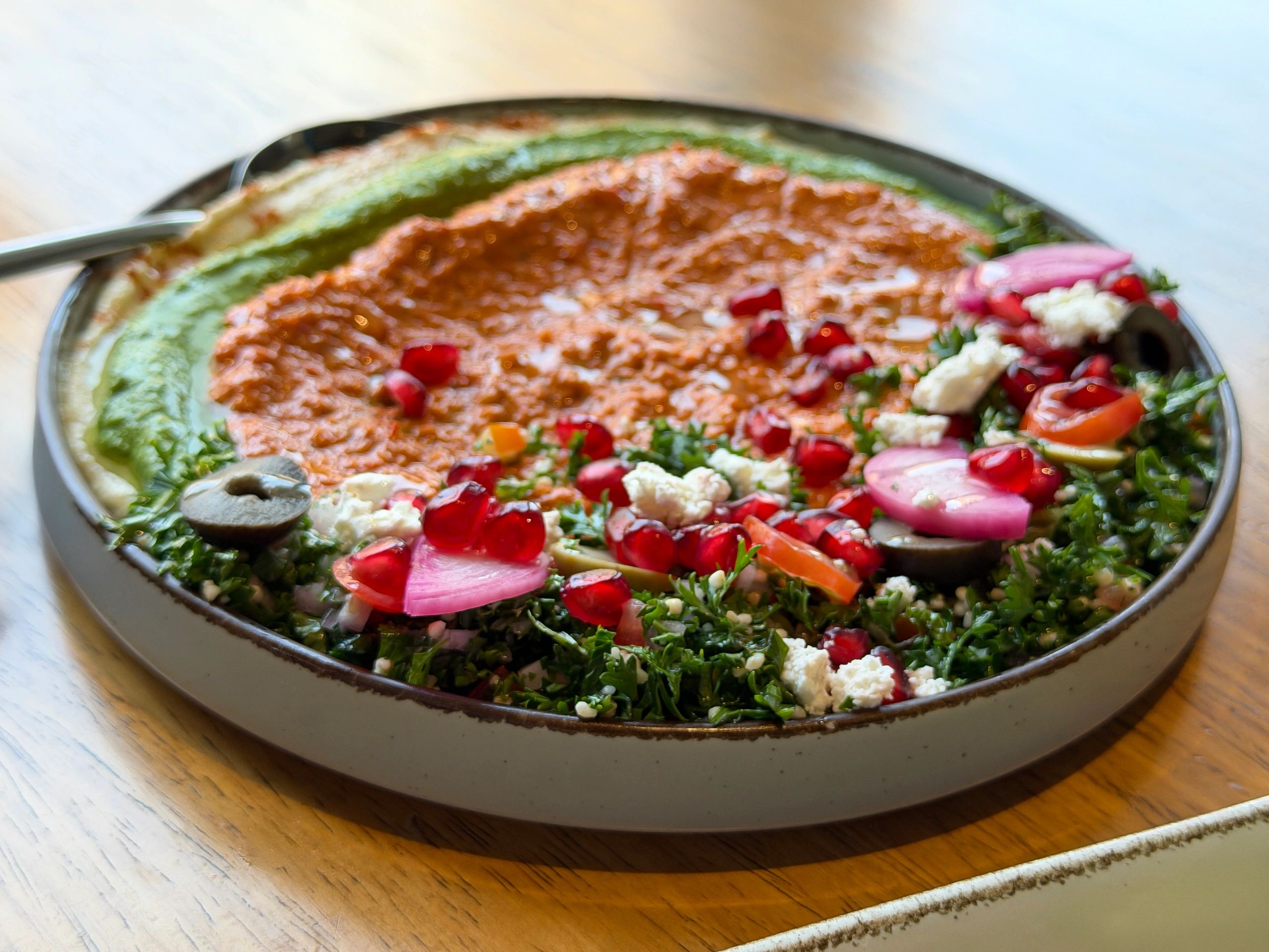 Image of Lebanese meza, bowl of houmous (chickpea dip), green pesto hummus and Muhammara (roasted red pepper walnut dip), pomegranate seeds, parsley, feta cheese, black and green olives, cherry tomato, pickled onion, metal spoon, elevated view